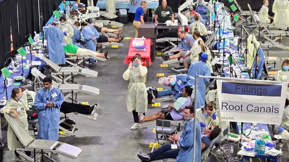 Inside an arena, rows of portable dental chairs line both sides of a wide concrete aisle. Patients recline in the chairs while volunteers in blue and yellow gowns and face masks treat them or stand by. A hanging sign on the right reads "Fillings" and "Root Canals" with a MOM-n-PA logo.