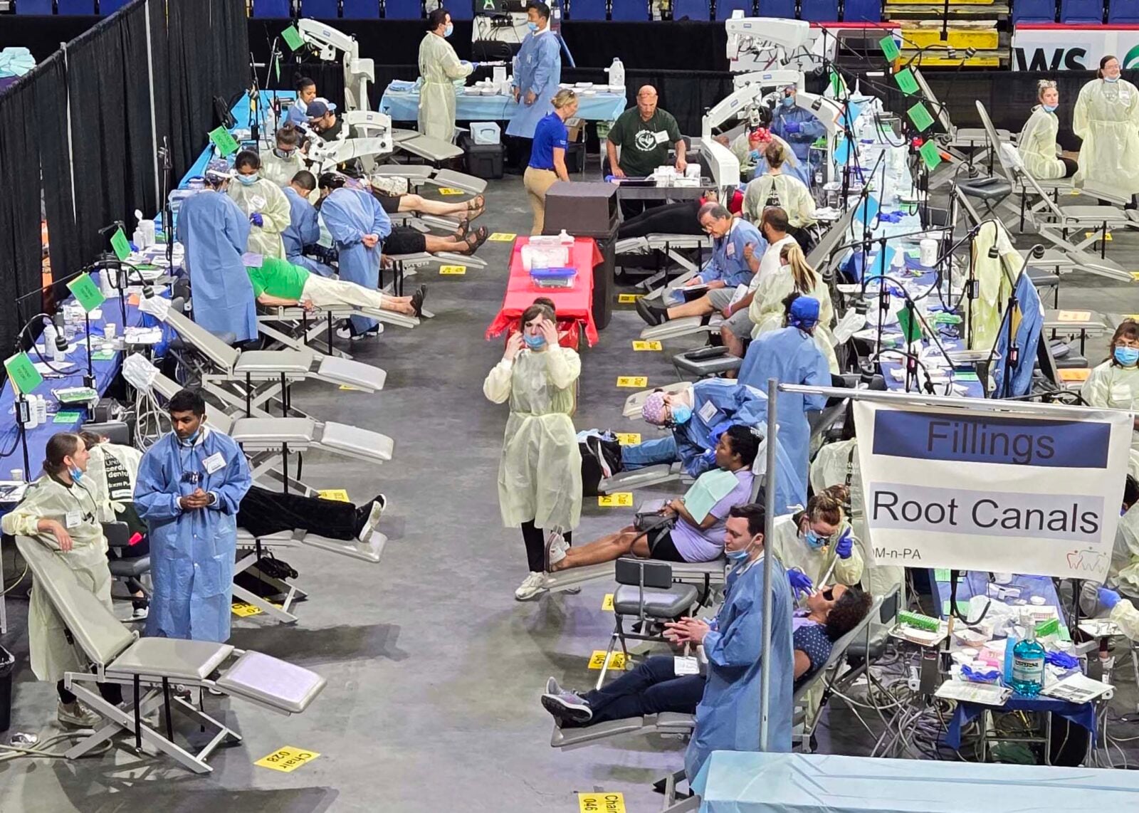 Inside an arena, rows of portable dental chairs line both sides of a wide concrete aisle. Patients recline in the chairs while volunteers in blue and yellow gowns and face masks treat them or stand by. A hanging sign on the right reads "Fillings" and "Root Canals" with a MOM-n-PA logo.