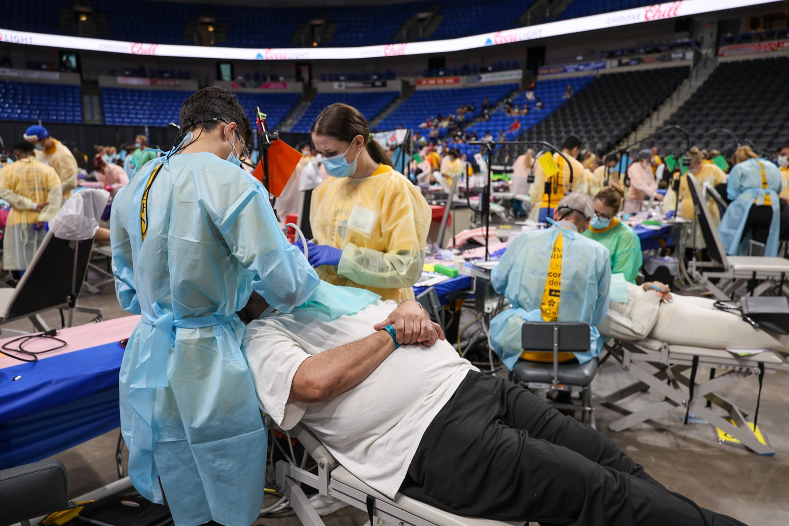 A wide view of an arena floor filled with rows of portable dental chairs. In the foreground, two masked volunteers in blue and yellow gowns treat a patient reclining in a chair. Dozens more volunteers and patients fill the rows behind them beneath empty blue stadium seats.