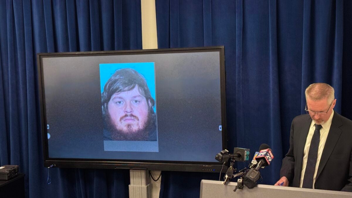 Lehigh County District Attorney Gavin Holihan stands at a podium with news microphones while a screen beside him displays a photo of Jack Pletz, a bearded young man with brown hair.