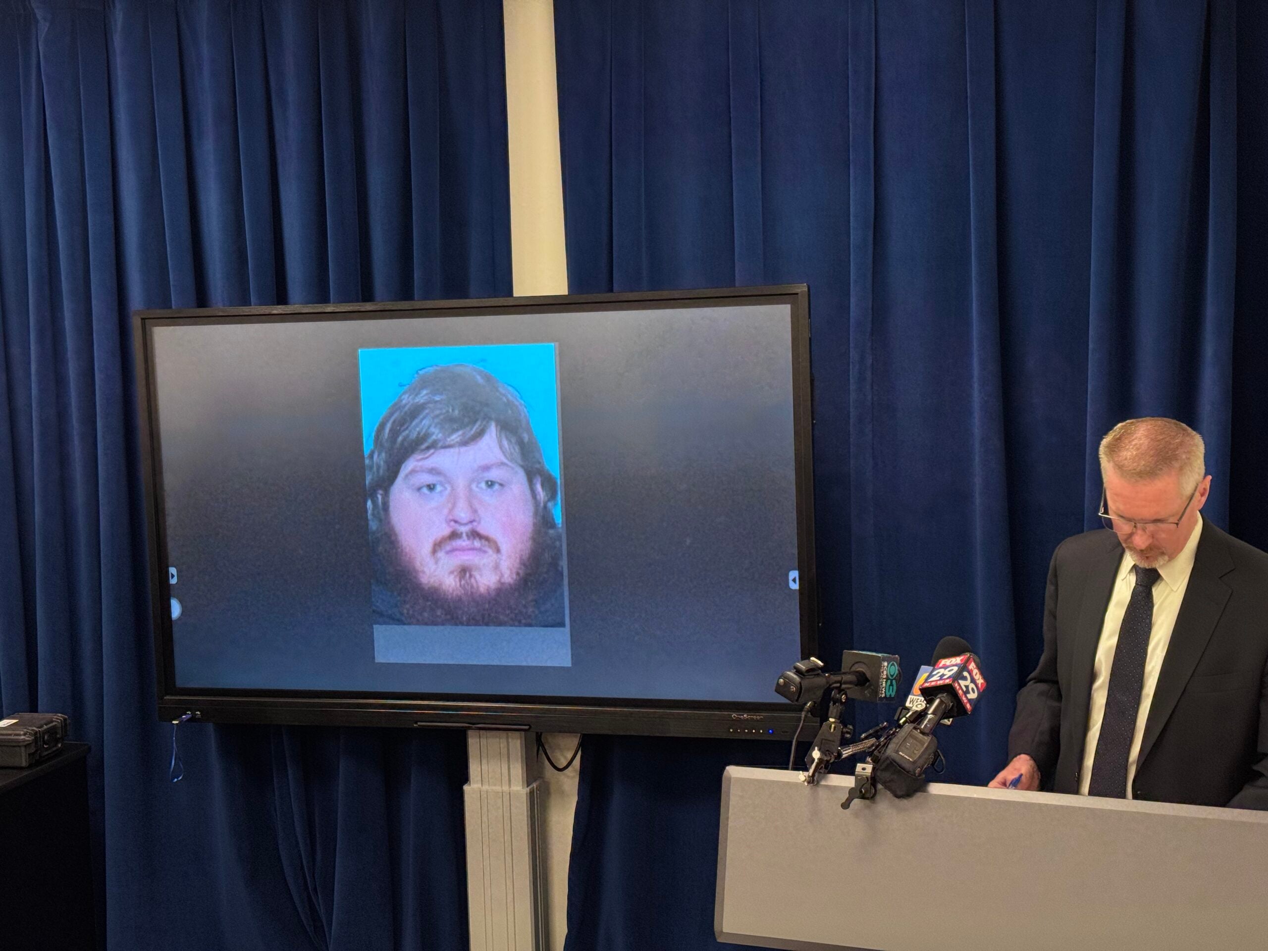 Lehigh County District Attorney Gavin Holihan stands at a podium with news microphones while a screen beside him displays a photo of Jack Pletz, a bearded young man with brown hair.
