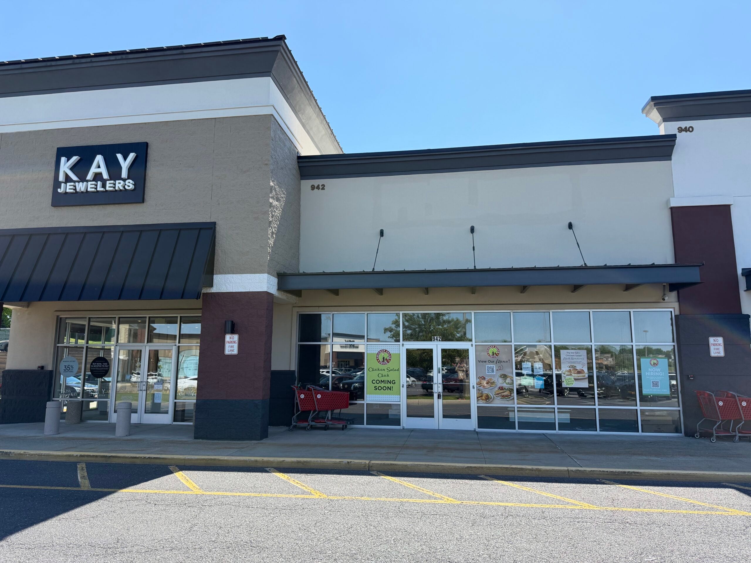 a storefront in a shopping center with a "chicken salad chick coming soon" sign in the window, next to a kay jewelers store, with shopping carts and a parking lot in the foreground