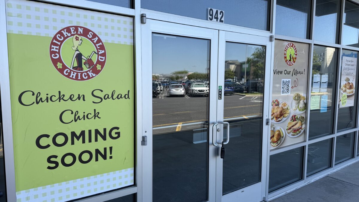 glass entrance doors marked 942 with a green "chicken salad chick coming soon" sign and a menu preview poster with photos of sandwiches and salads taped to the window