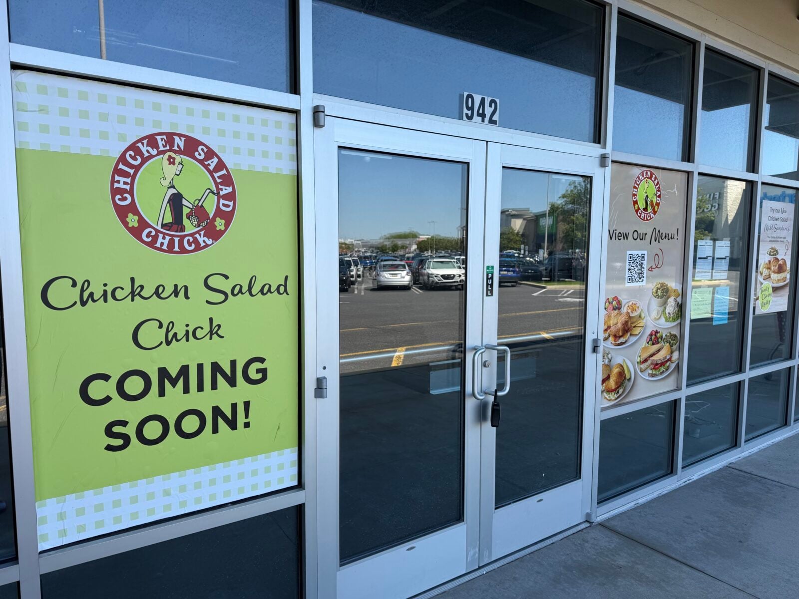 glass entrance doors marked 942 with a green "chicken salad chick coming soon" sign and a menu preview poster with photos of sandwiches and salads taped to the window