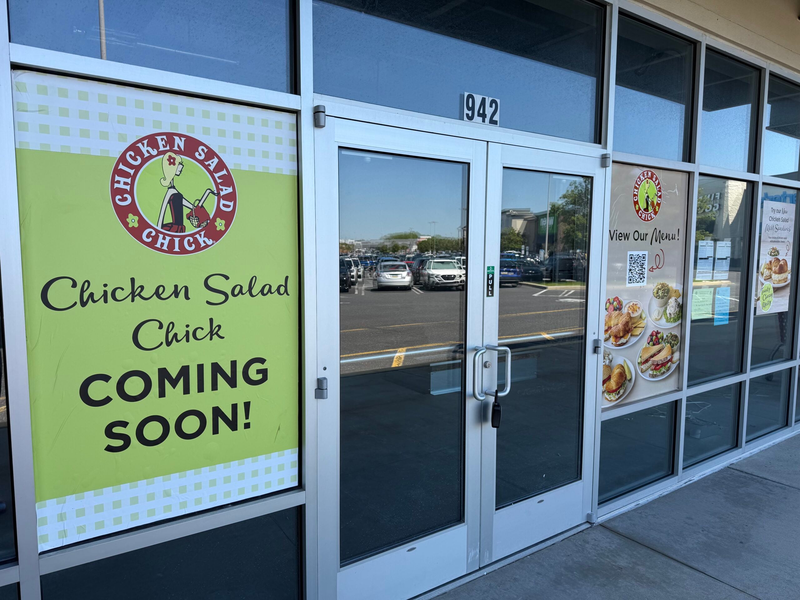 glass entrance doors marked 942 with a green "chicken salad chick coming soon" sign and a menu preview poster with photos of sandwiches and salads taped to the window