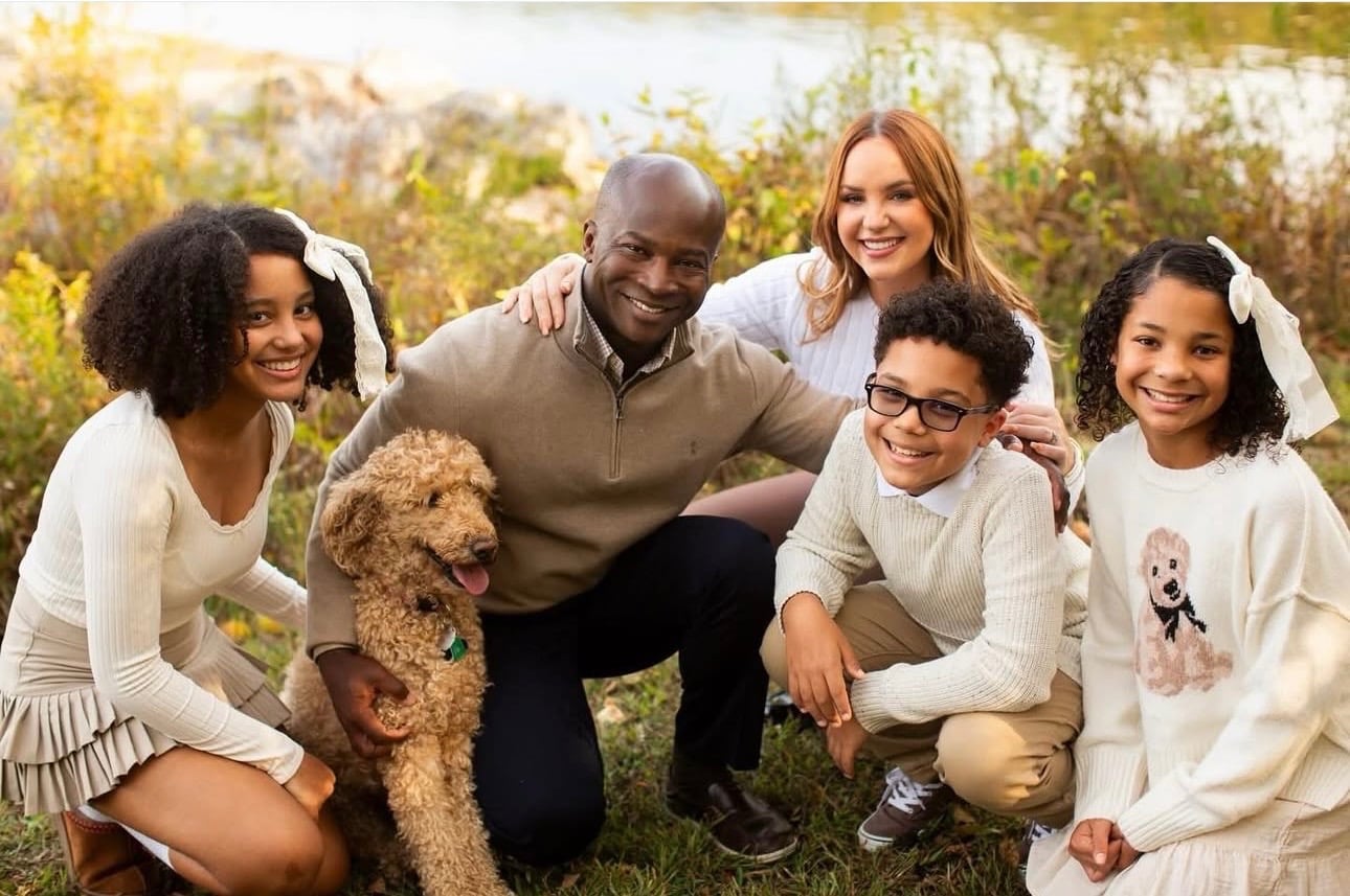 a family of two adults and three children kneeling on grass beside a lake with a curly-haired tan dog, all dressed in cream and tan clothing