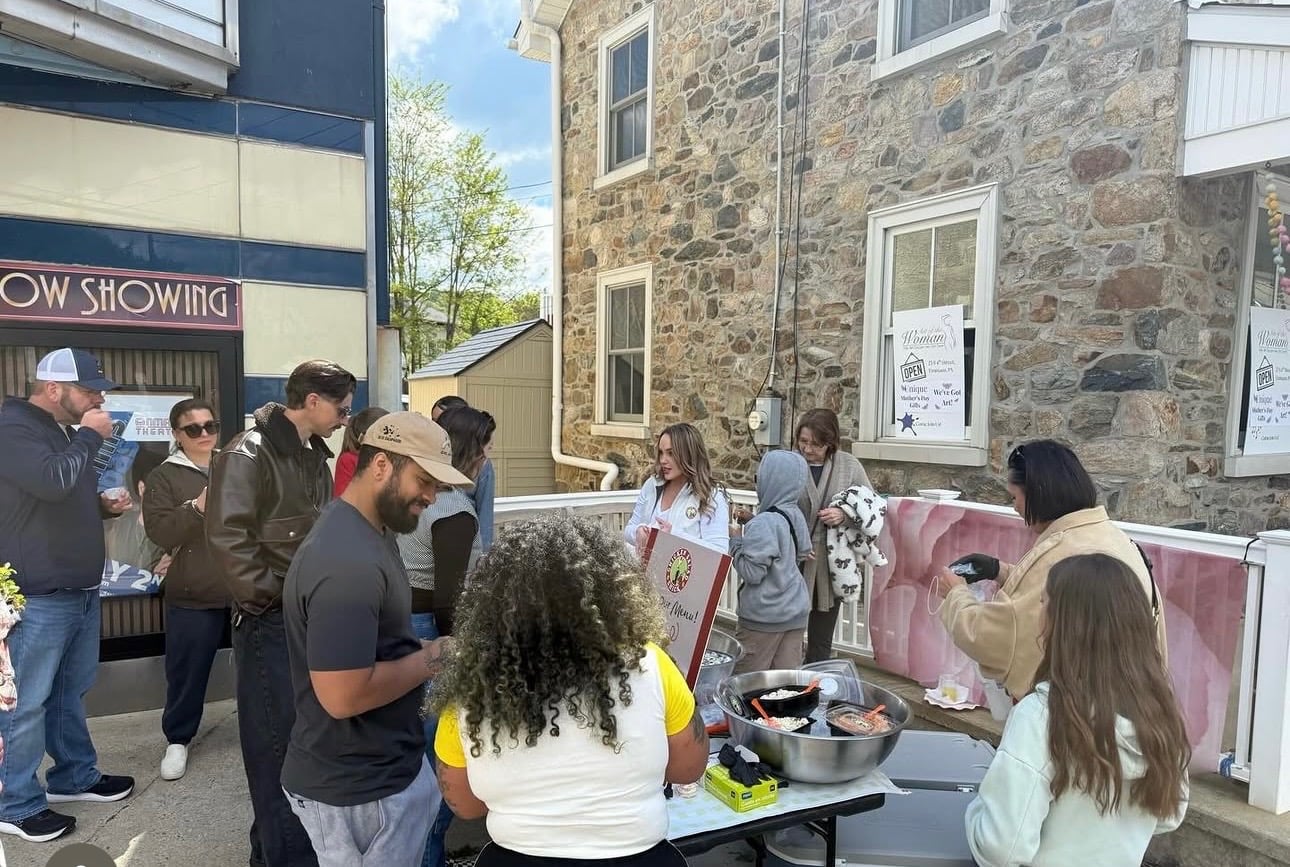 a woman in a white chicken salad chick jacket serving food from metal bowls at an outdoor table as people gather around, in front of a stone building and a movie theater marquee