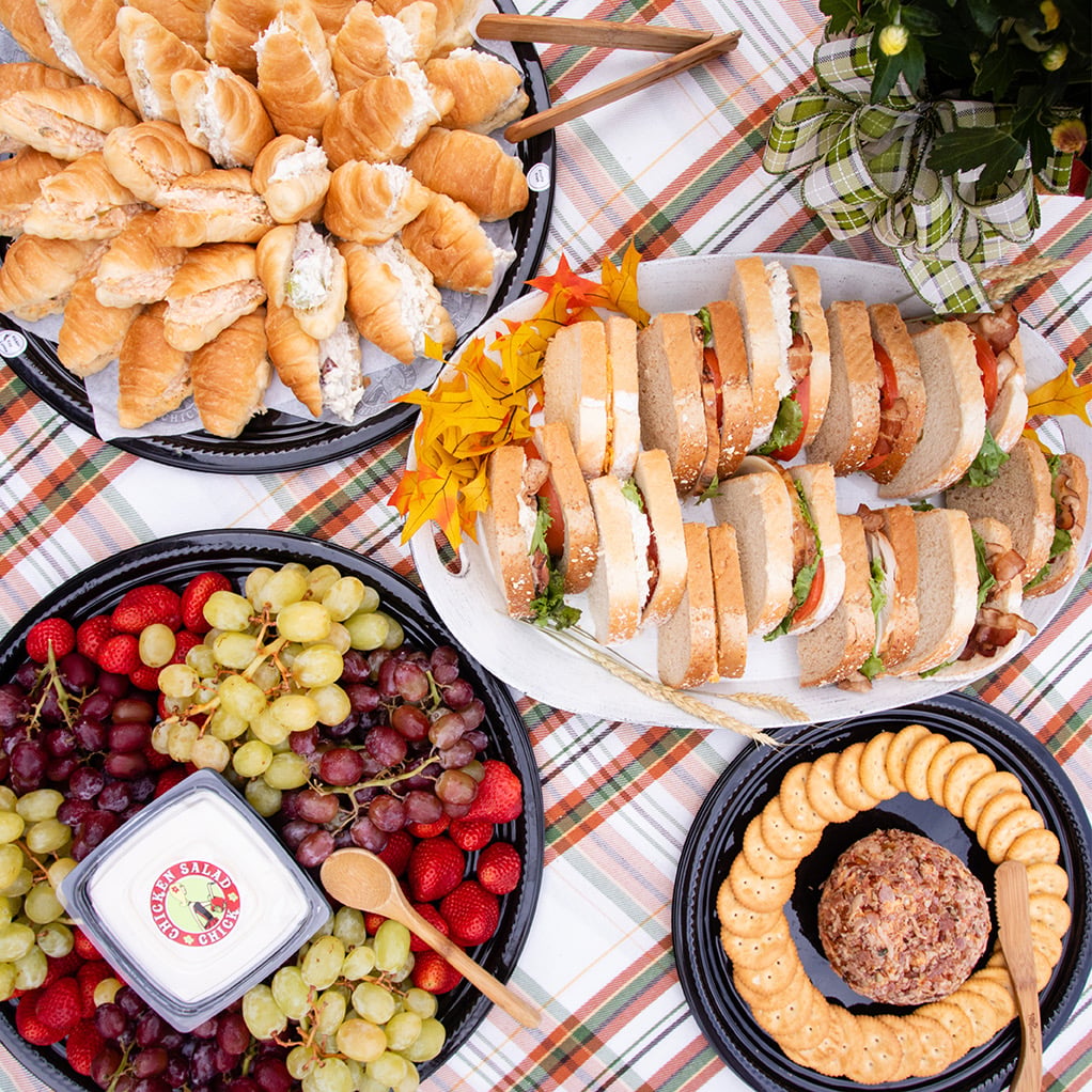an overhead view of catering platters on a plaid cloth with mini croissant sandwiches, club sandwiches, a tray of grapes and strawberries with dip, and a cheese ball with round crackers