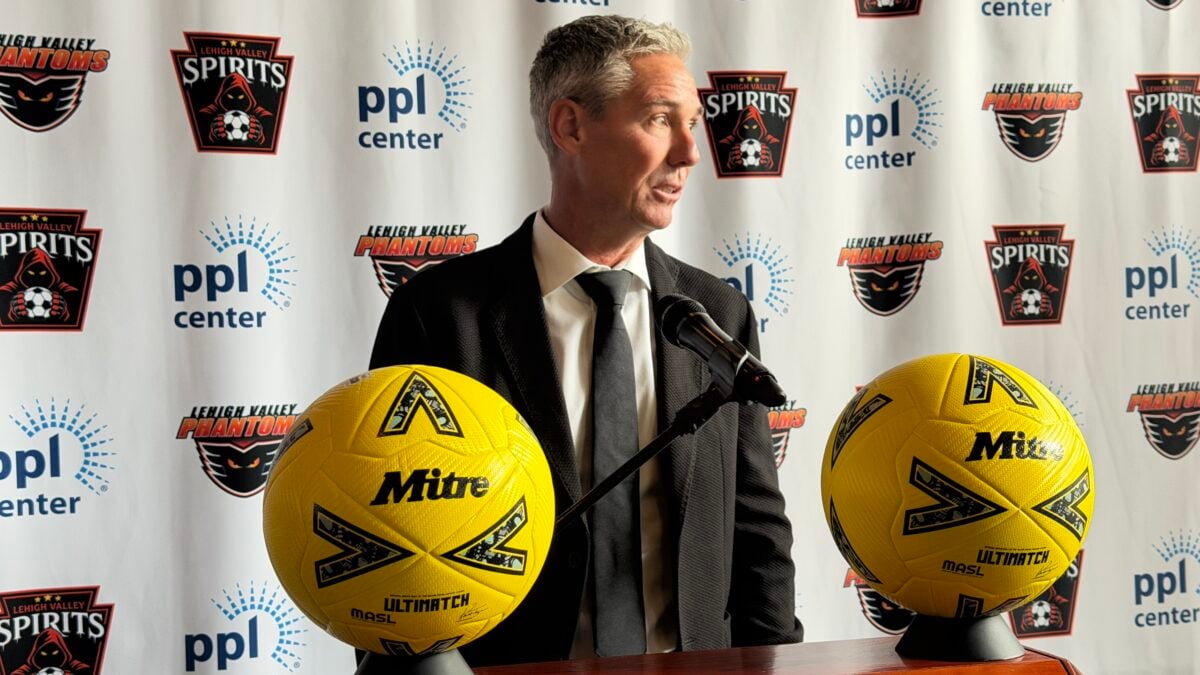 Lehigh Valley Spirits Sporting Director Giuliano Oliviero, a gray-haired man in a dark suit and tie, speaks at a podium with a microphone, flanked by two yellow Mitre soccer balls, in front of a backdrop repeating the Lehigh Valley Spirits, Lehigh Valley Phantoms and PPL Center logos.