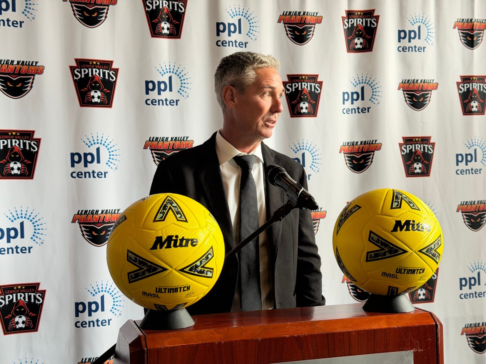 Lehigh Valley Spirits Sporting Director Giuliano Oliviero, a gray-haired man in a dark suit and tie, speaks at a podium with a microphone, flanked by two yellow Mitre soccer balls, in front of a backdrop repeating the Lehigh Valley Spirits, Lehigh Valley Phantoms and PPL Center logos.