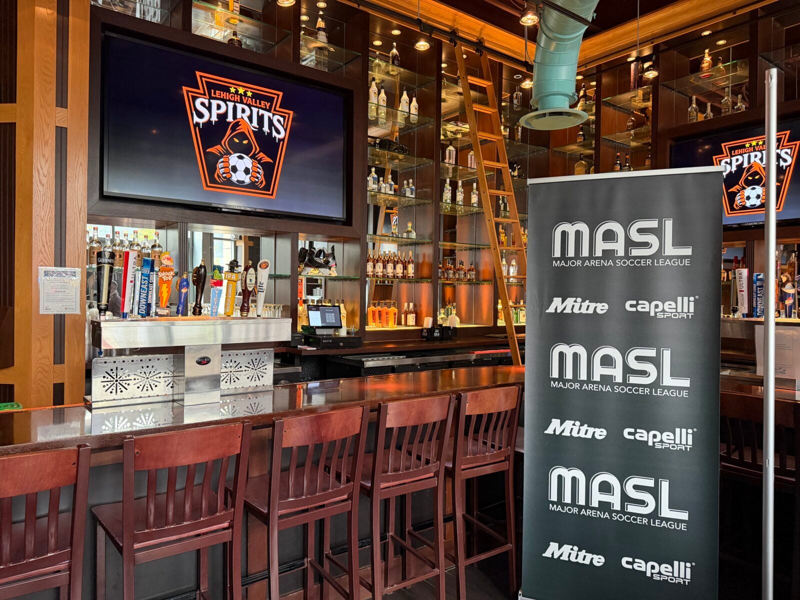 A bar at Chickie's & Pete's at PPL Center, where a wall-mounted screen shows the Lehigh Valley Spirits logo and a freestanding banner displays the Major Arena Soccer League logo alongside Mitre and Capelli Sport branding.