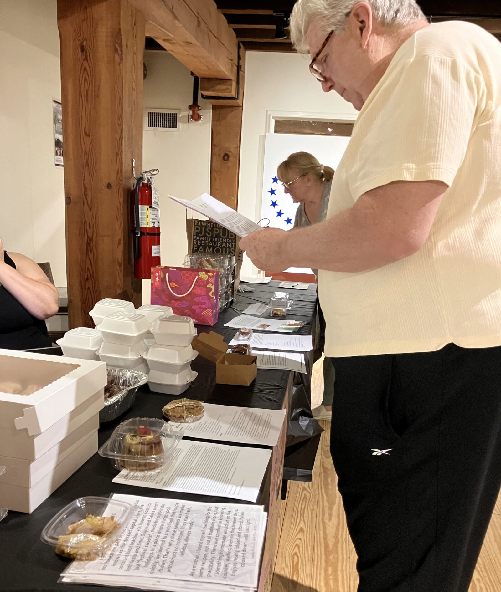 richard groman jr., of the family behind the longtime groman's bakery, reads a printed recipe sheet while judging the first round of the baked into bethlehem baking challenge, standing over a long black-draped table lined with home-baked desserts in clear and foam containers; a second judge reviews papers in the background of the timber-framed room