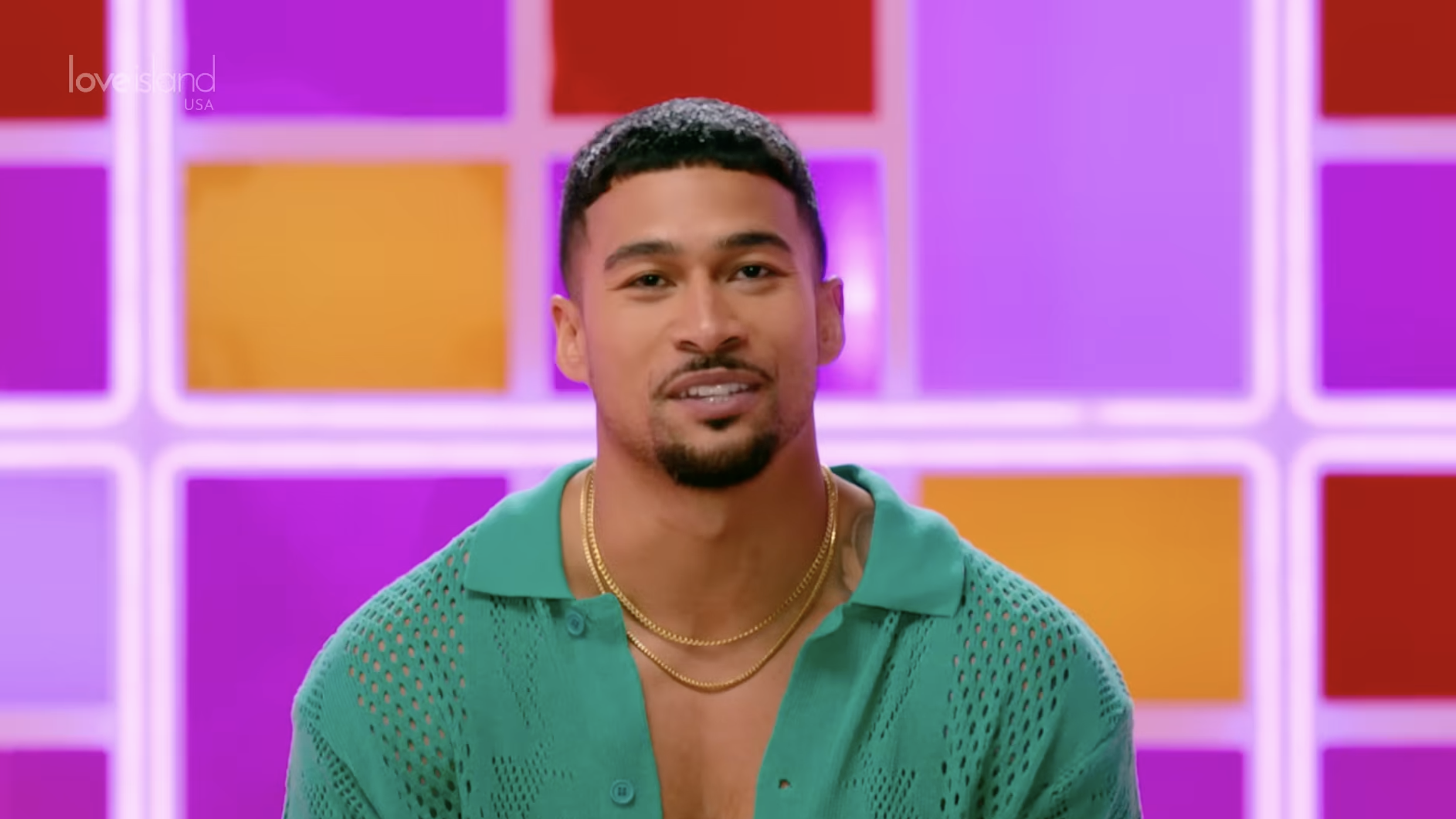 Sean Reifel, a Bethlehem police officer cast on "Love Island USA" season 8, smiles in an open green knit polo shirt and two gold chains, against a backlit pink, purple, orange and red grid wall with the "Love Island USA" logo in the upper left.