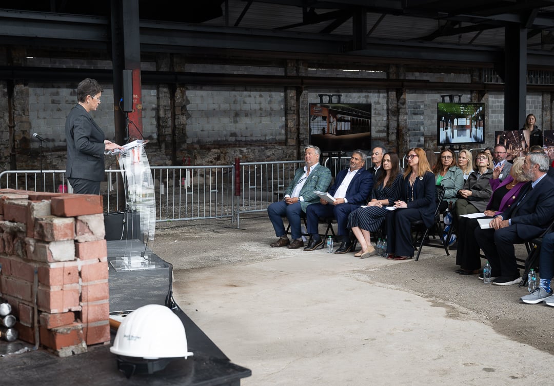 a woman in a dark suit speaks at a clear podium inside the unfinished brick turn & grind shop building, with a stack of bricks and a white hard hat in the foreground and rows of seated attendees facing her