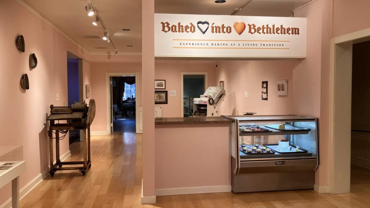 a museum gallery with pale pink walls and light wood floors, where a white sign reading "baked into bethlehem" hangs above a mock bakery counter with a glass display case of small cakes and pastries; an old dough-rolling machine sits on a cart against the wall to the left, with open doorways leading to other rooms