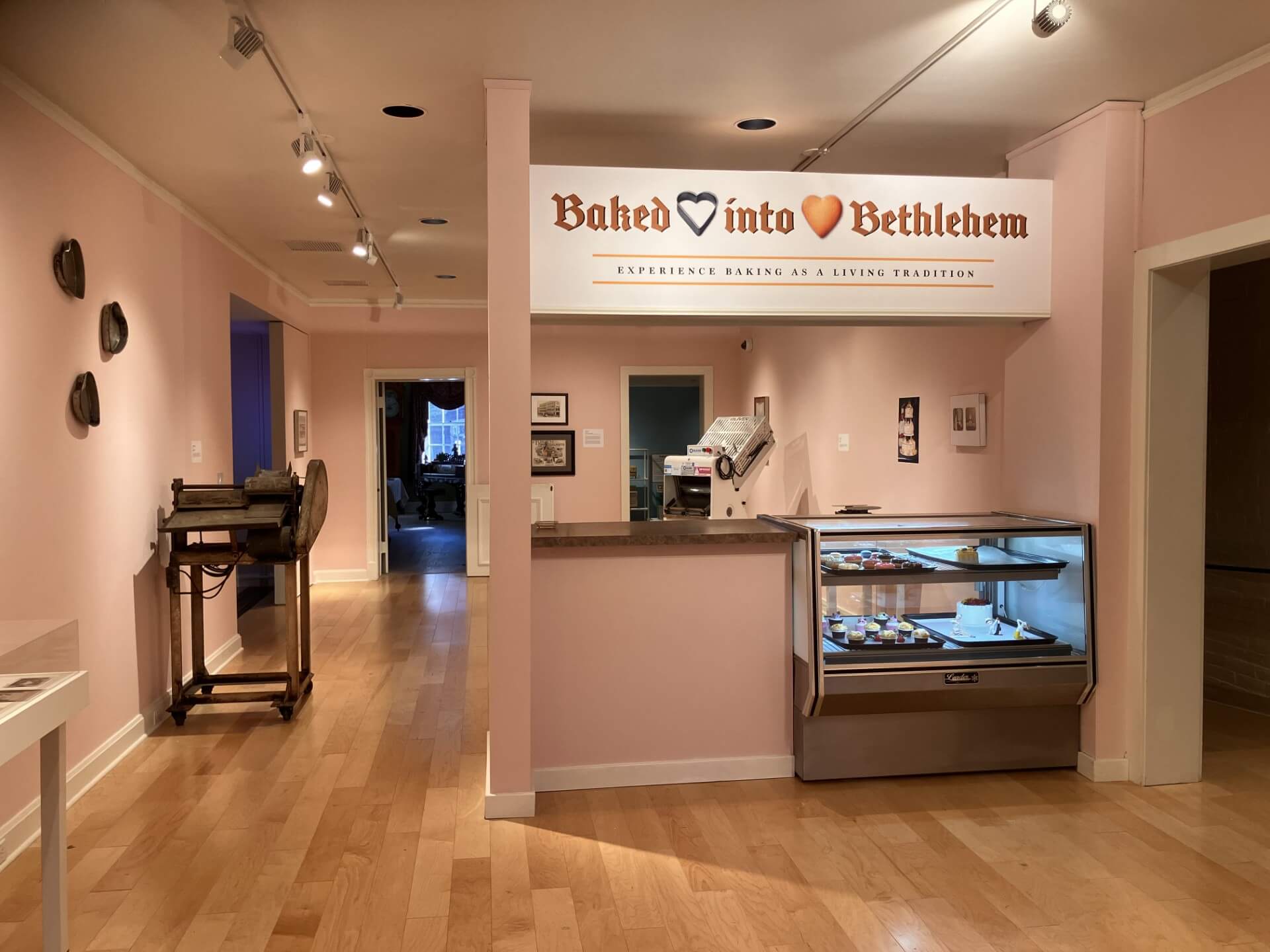 a museum gallery with pale pink walls and light wood floors, where a white sign reading "baked into bethlehem" hangs above a mock bakery counter with a glass display case of small cakes and pastries; an old dough-rolling machine sits on a cart against the wall to the left, with open doorways leading to other rooms