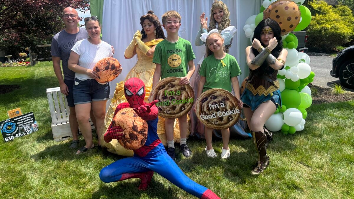 A boy stands outdoors with his family and costumed performers dressed as Spider-Man, Wonder Woman, and Disney princesses, several holding large cookie-shaped props, in front of a green "Be a Good Cookie" banner and a green-and-white balloon arch.