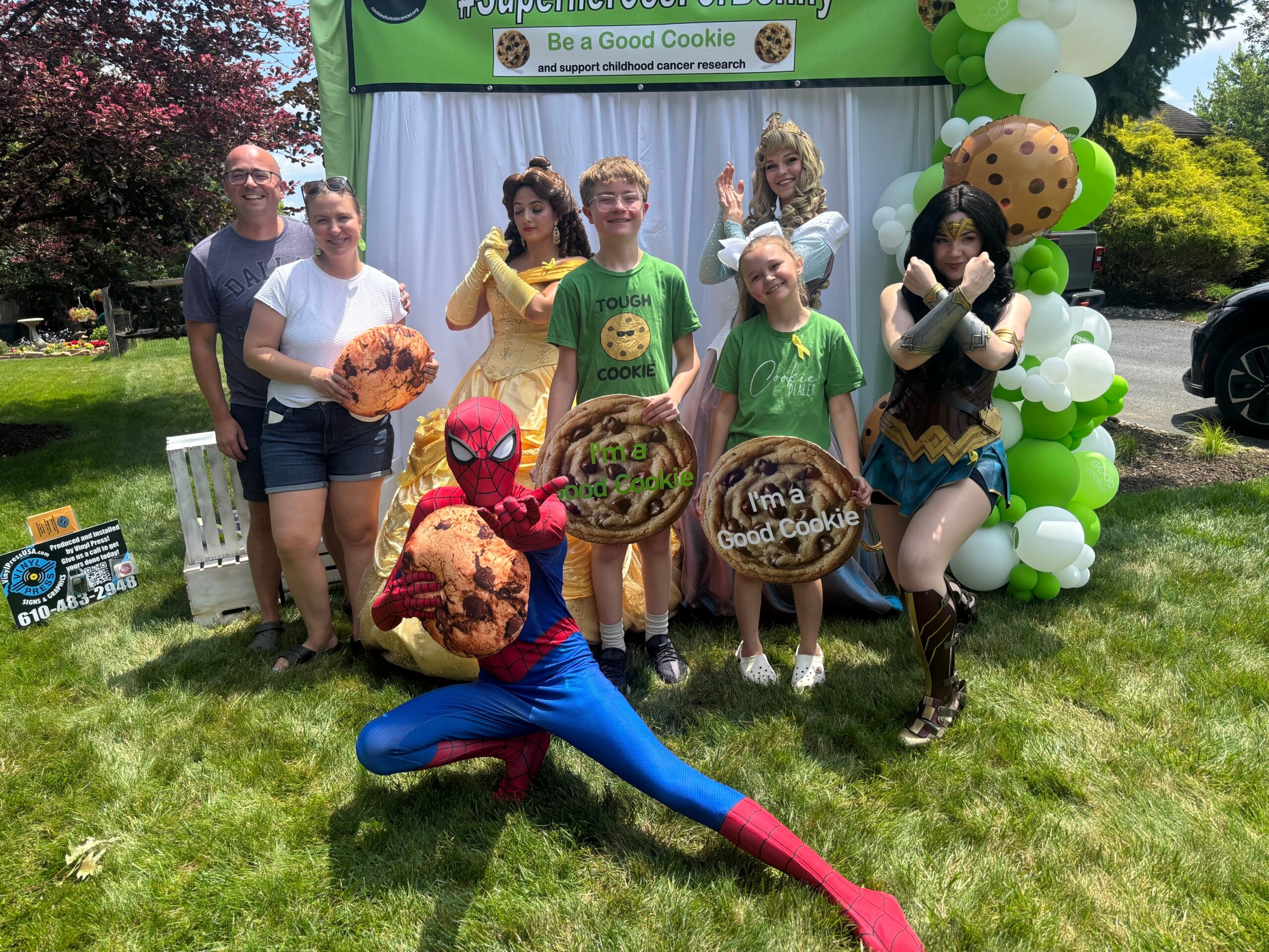 A boy stands outdoors with his family and costumed performers dressed as Spider-Man, Wonder Woman, and Disney princesses, several holding large cookie-shaped props, in front of a green "Be a Good Cookie" banner and a green-and-white balloon arch.