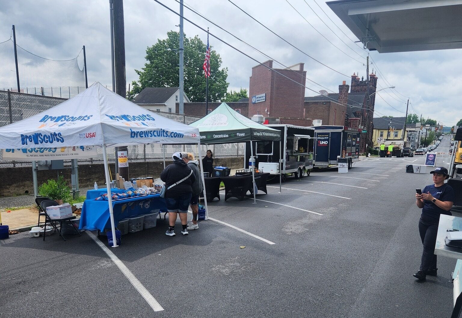 vendor tents and food trailers set up along a closed street, with two people browsing a tent table and a worker standing nearby