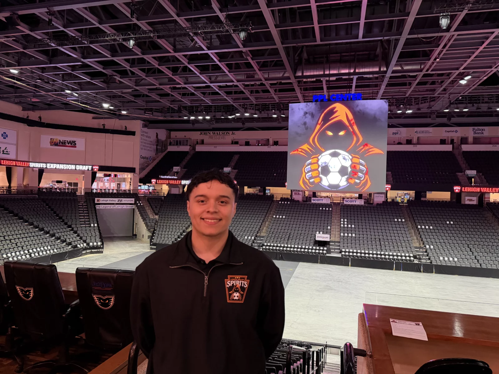 forward jairo guevara wears a black lehigh valley spirits quarter-zip and stands inside ppl center in front of empty seats, with the team's orange hooded figure logo displayed on the video board behind him