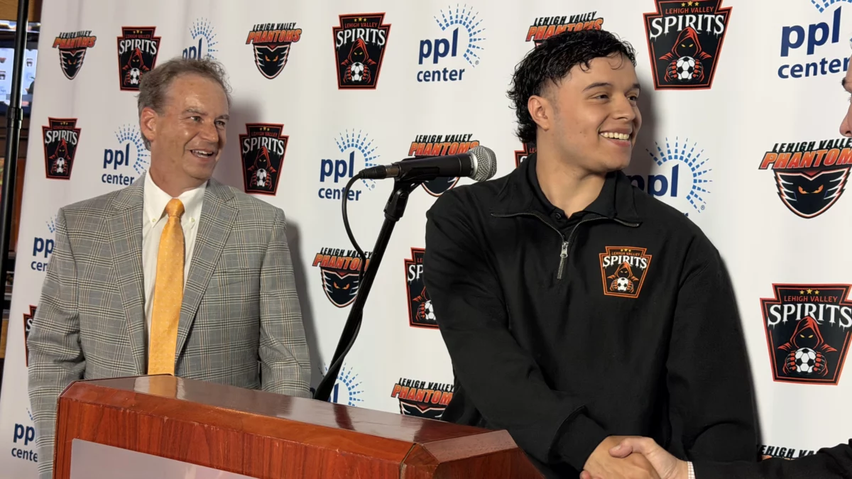 lehigh valley spirits co-owner rob brooks stands at a podium on the left as forward jairo guevara, wearing a black spirits quarter-zip, shakes hands with co-owner jim brooks in front of a ppl center and lehigh valley spirits logo backdrop