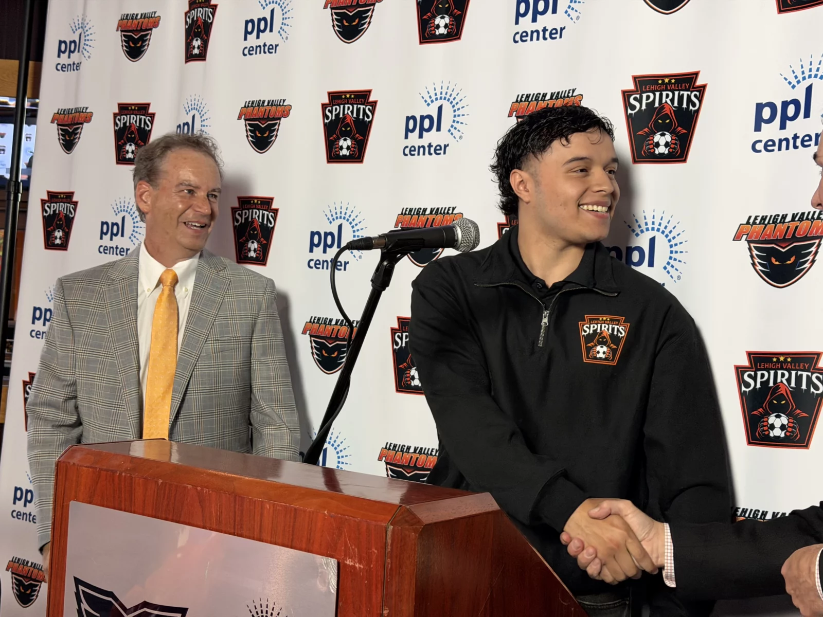 lehigh valley spirits co-owner rob brooks stands at a podium on the left as forward jairo guevara, wearing a black spirits quarter-zip, shakes hands with co-owner jim brooks in front of a ppl center and lehigh valley spirits logo backdrop