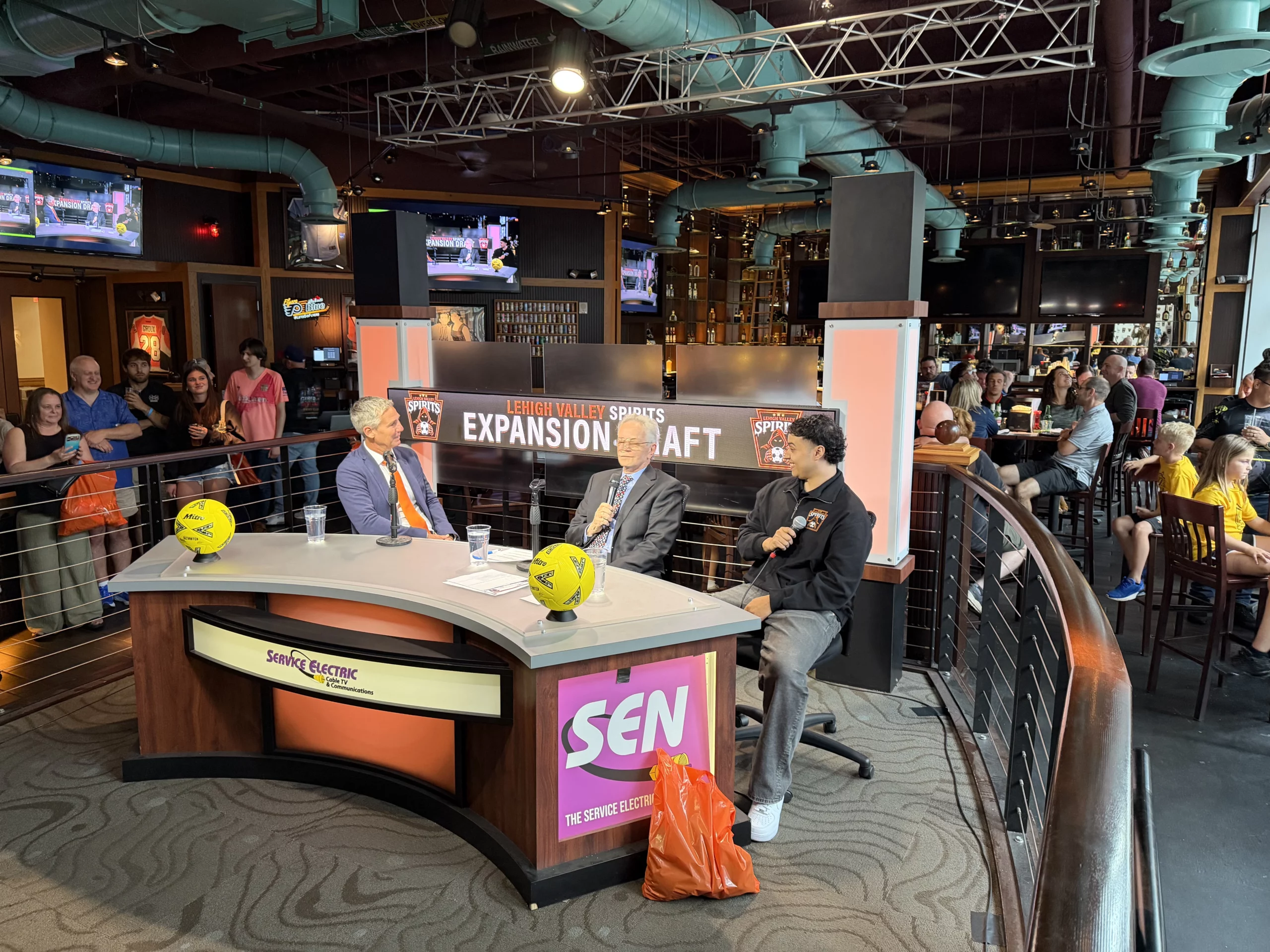 from left, lehigh valley spirits sporting director giuliano oliviero, masl president of media and communications jp dellacamera and forward jairo guevara sit at a broadcast desk with yellow mitre soccer balls in front of a lehigh valley spirits expansion draft backdrop inside chickie's and pete's