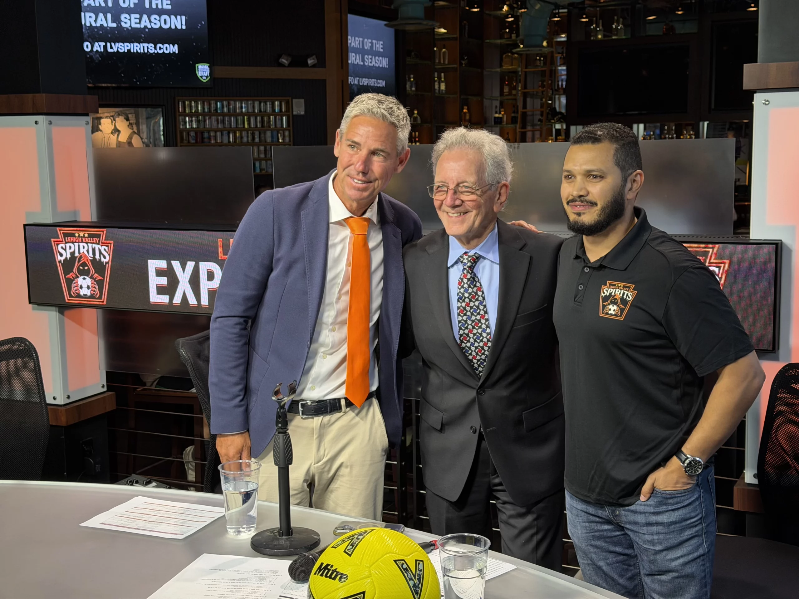 from left, lehigh valley spirits sporting director giuliano oliviero, masl president of media and communications jp dellacamera and spirits associate head coach jeremy ortiz stand together at the broadcast desk in front of a lehigh valley spirits expansion draft backdrop inside chickie's and pete's
