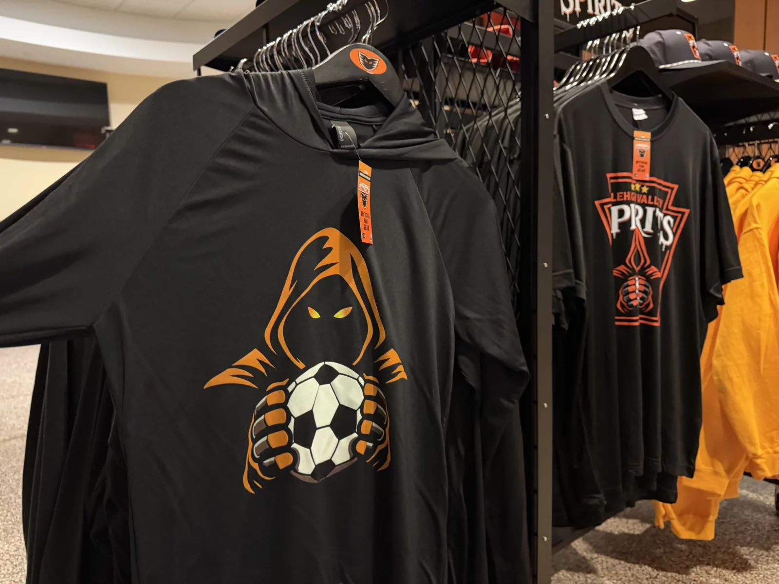 lehigh valley spirits black and yellow t-shirts featuring the team's orange hooded figure logo holding a soccer ball, hanging on a retail rack at chickie's and pete's Image 2 (broadcast desk): from left, lehigh valley spirits sporting director giuliano oliviero, masl president of media and communications jp dellacamera and forward jairo guevara sit at a broadcast desk with yellow mitre soccer balls in front of a lehigh valley spirits expansion draft backdrop inside chickie's and pete's