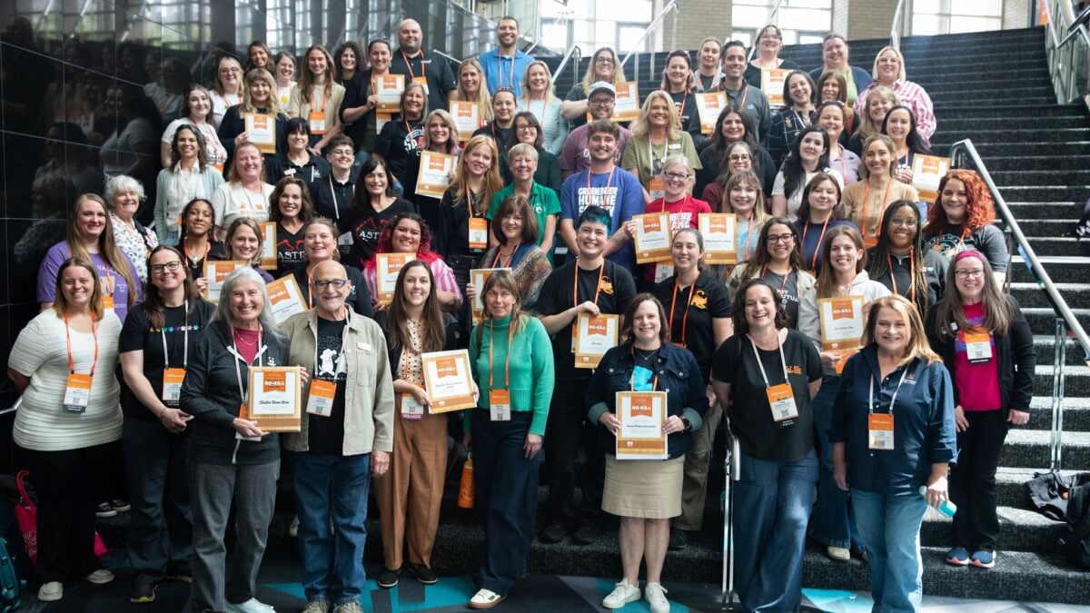 Members of the Lehigh Valley Humane Society stand with representatives from other shelters in a large group of several dozen people on an interior staircase, smiling toward the camera. Many wear orange lanyards and hold framed and paper certificates labeled "No-Kill." Some shirts and badges identify various animal shelters and humane societies. Tall windows and a patterned teal-and-gray floor are visible in the background.