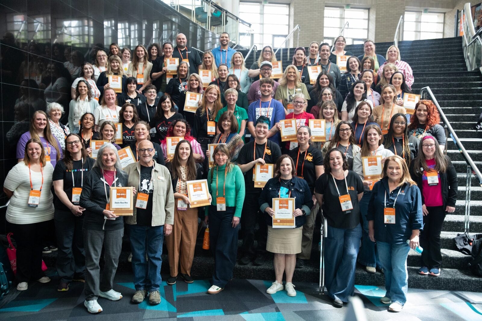 Members of the Lehigh Valley Humane Society stand with representatives from other shelters in a large group of several dozen people on an interior staircase, smiling toward the camera. Many wear orange lanyards and hold framed and paper certificates labeled "No-Kill." Some shirts and badges identify various animal shelters and humane societies. Tall windows and a patterned teal-and-gray floor are visible in the background.