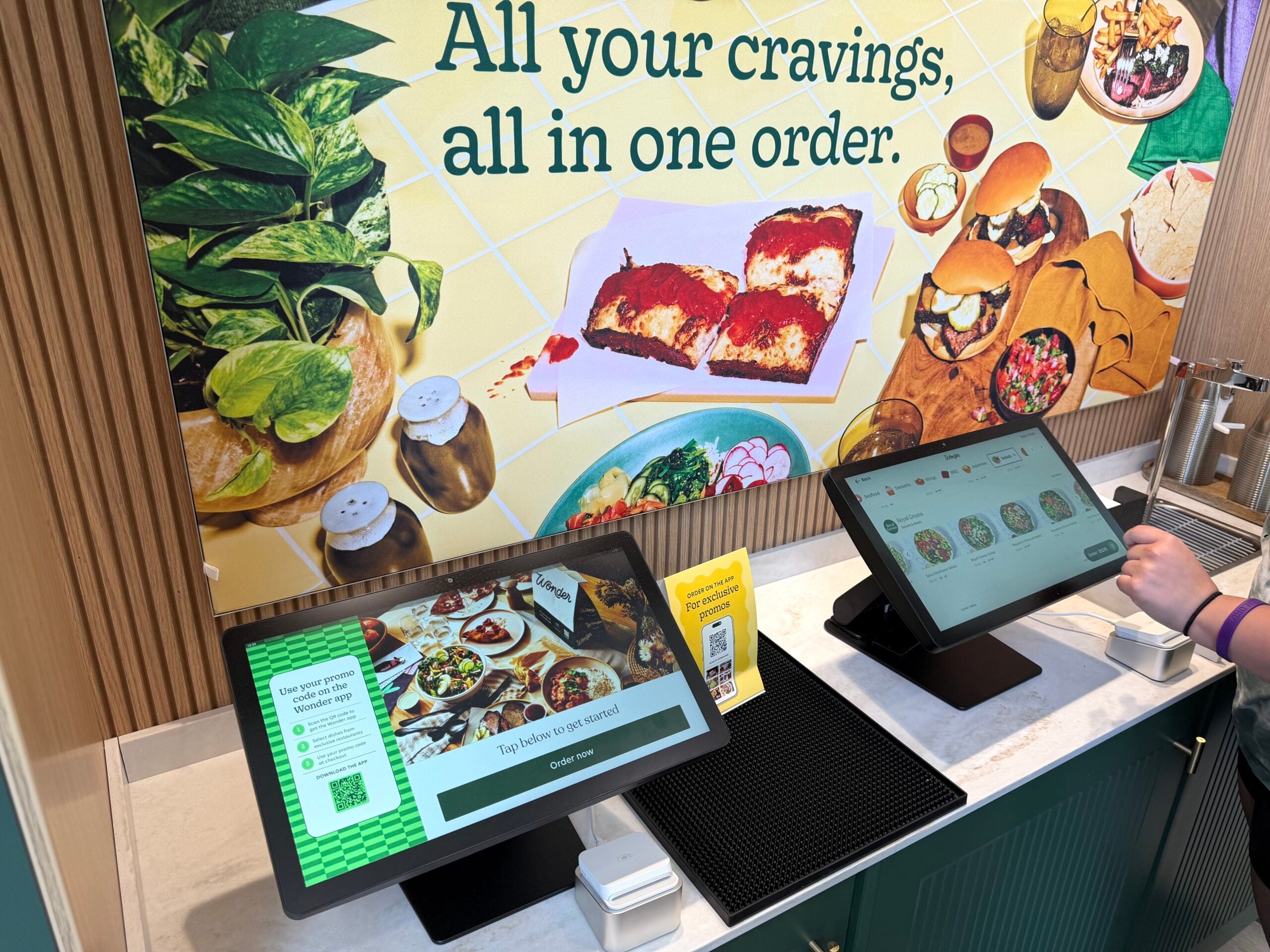 Two self-service ordering kiosks on a counter, one screen touched by a hand. Food mural behind.