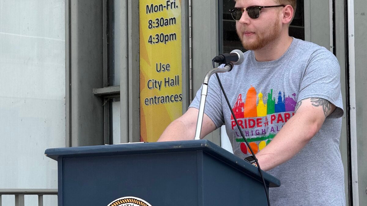 A man with a beard and sunglasses speaks at a podium, wearing a gray Pride t-shirt.