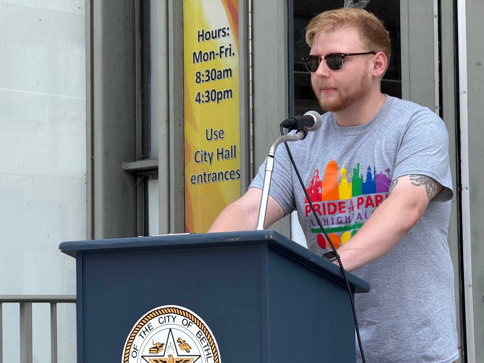 A man with a beard and sunglasses speaks at a podium, wearing a gray Pride t-shirt.