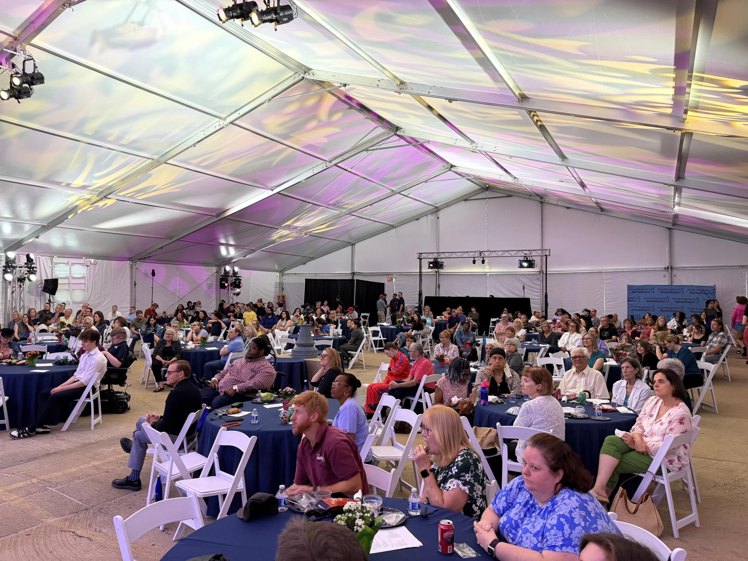 A large audience sits at round tables inside a white event tent with colorful light patterns on the ceiling.