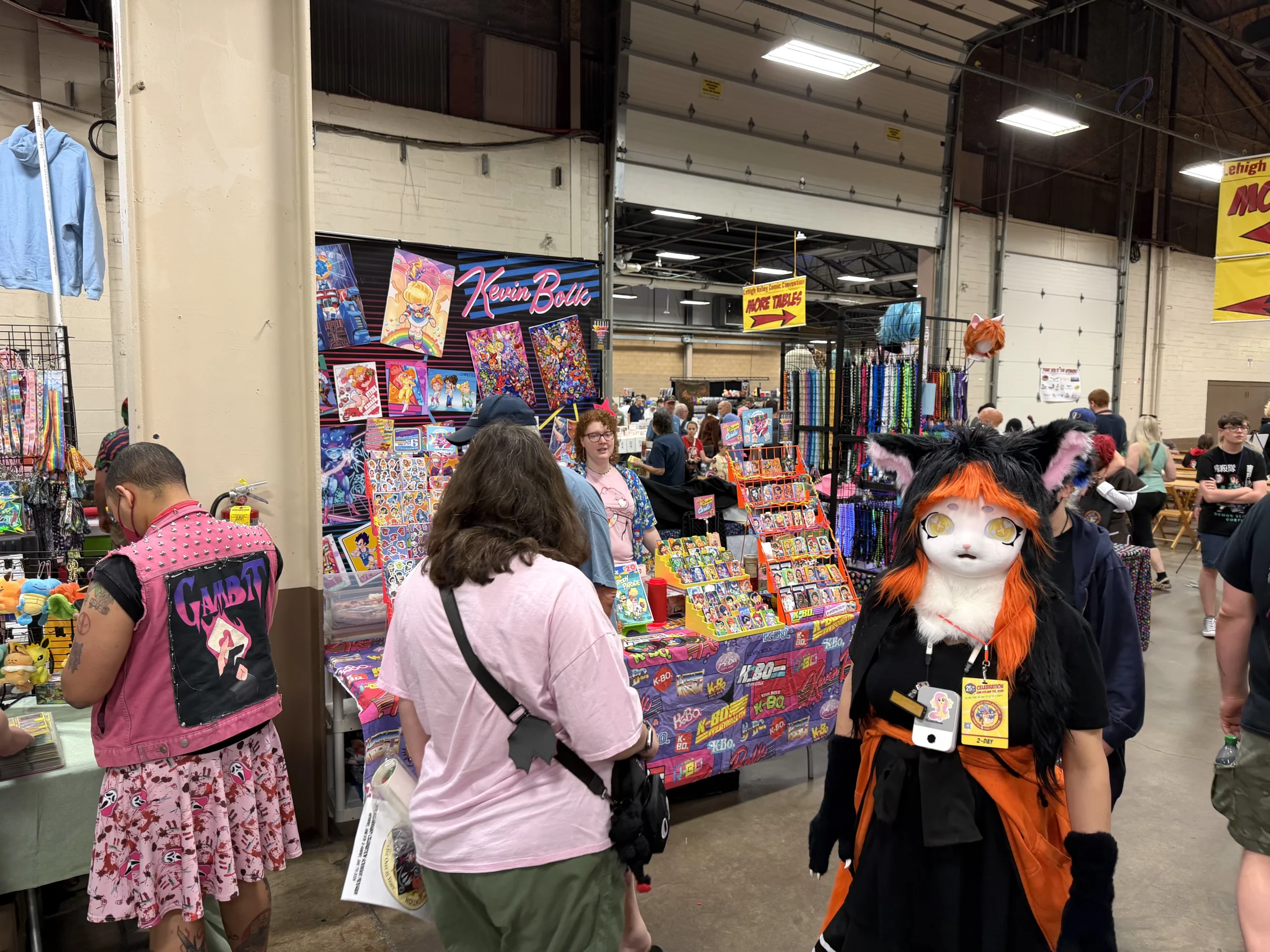 A person in a black and orange cat costume stands in a busy convention hall. Other attendees browse.