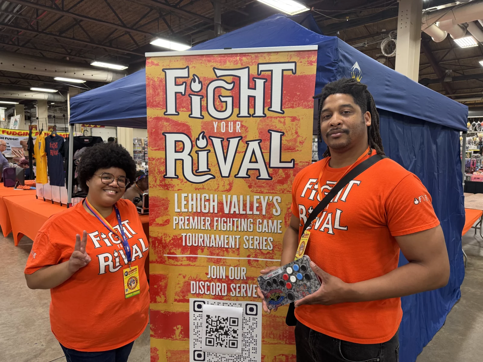 Two people in orange shirts stand by a "Fight Your Rival" banner, one holding a game controller.