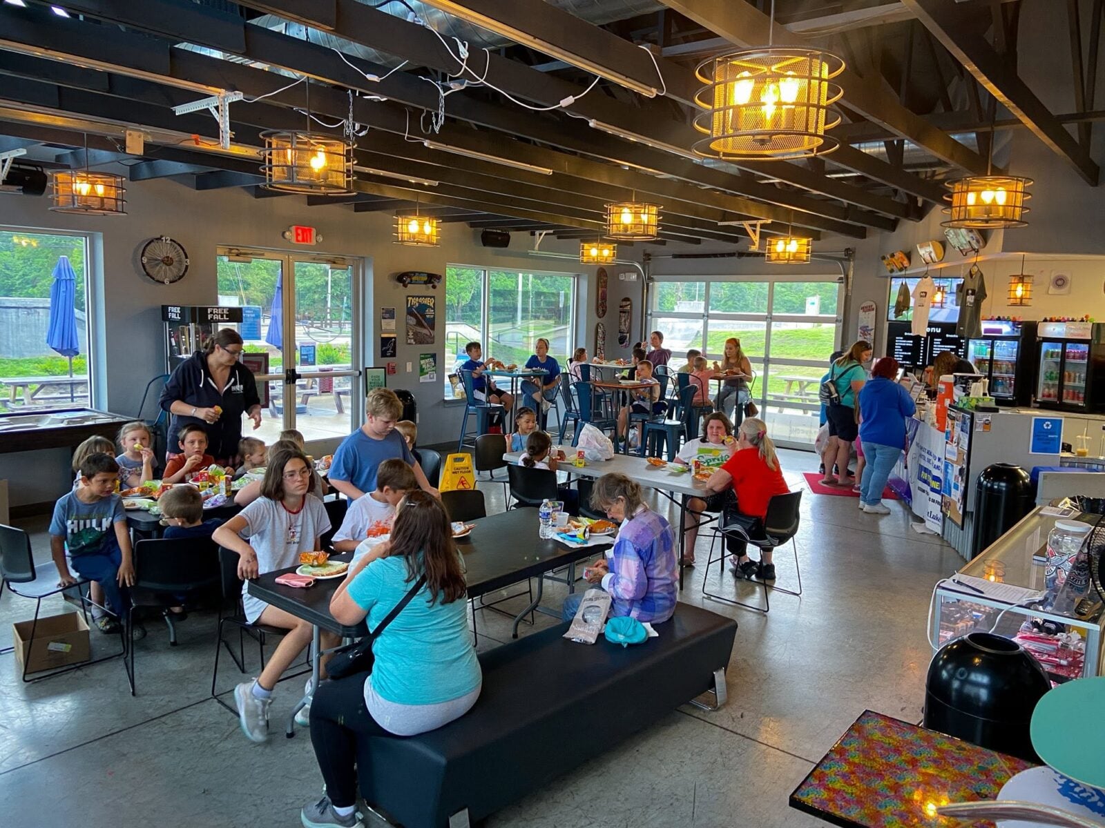 Children and adults eat meals at tables inside a large, brightly lit recreational facility.