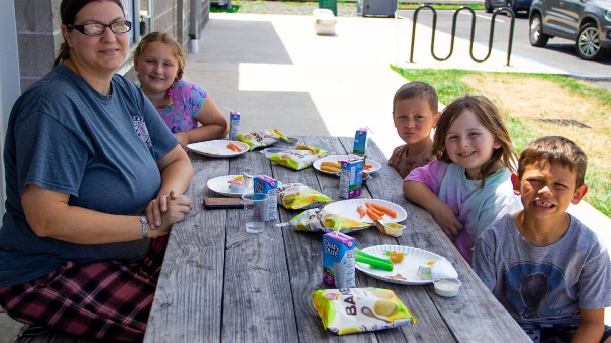 A woman and four children sit at a picnic table with plates of food and drinks.