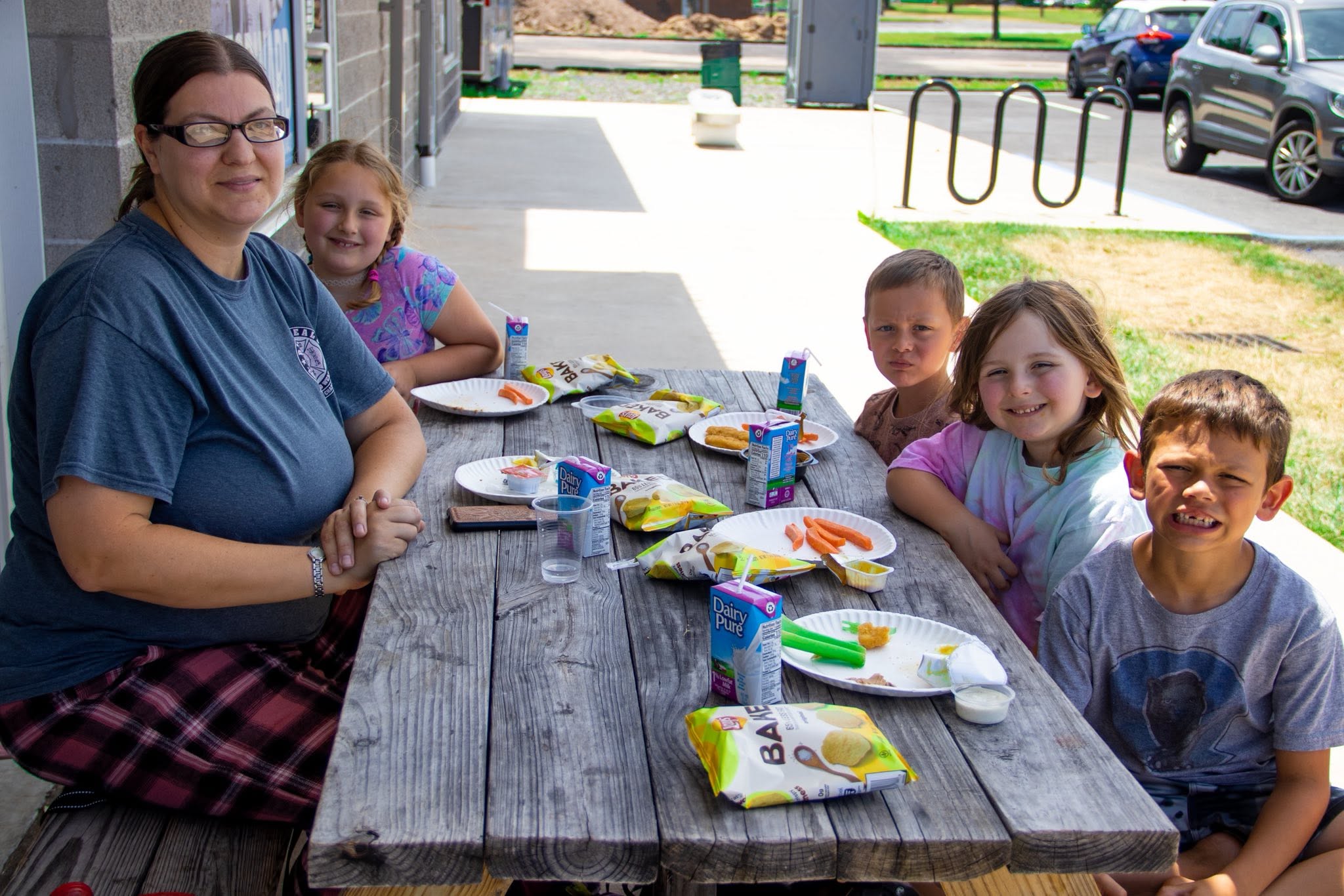 A woman and four children sit at a picnic table with plates of food and drinks.