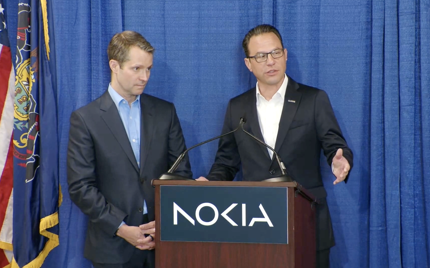 Two men in suits stand at a podium with the Nokia logo, one speaking.