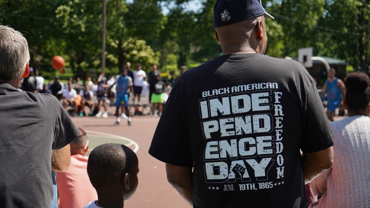 People watch an outdoor basketball game; one person wears a shirt saying 'Black American Independence Day'.