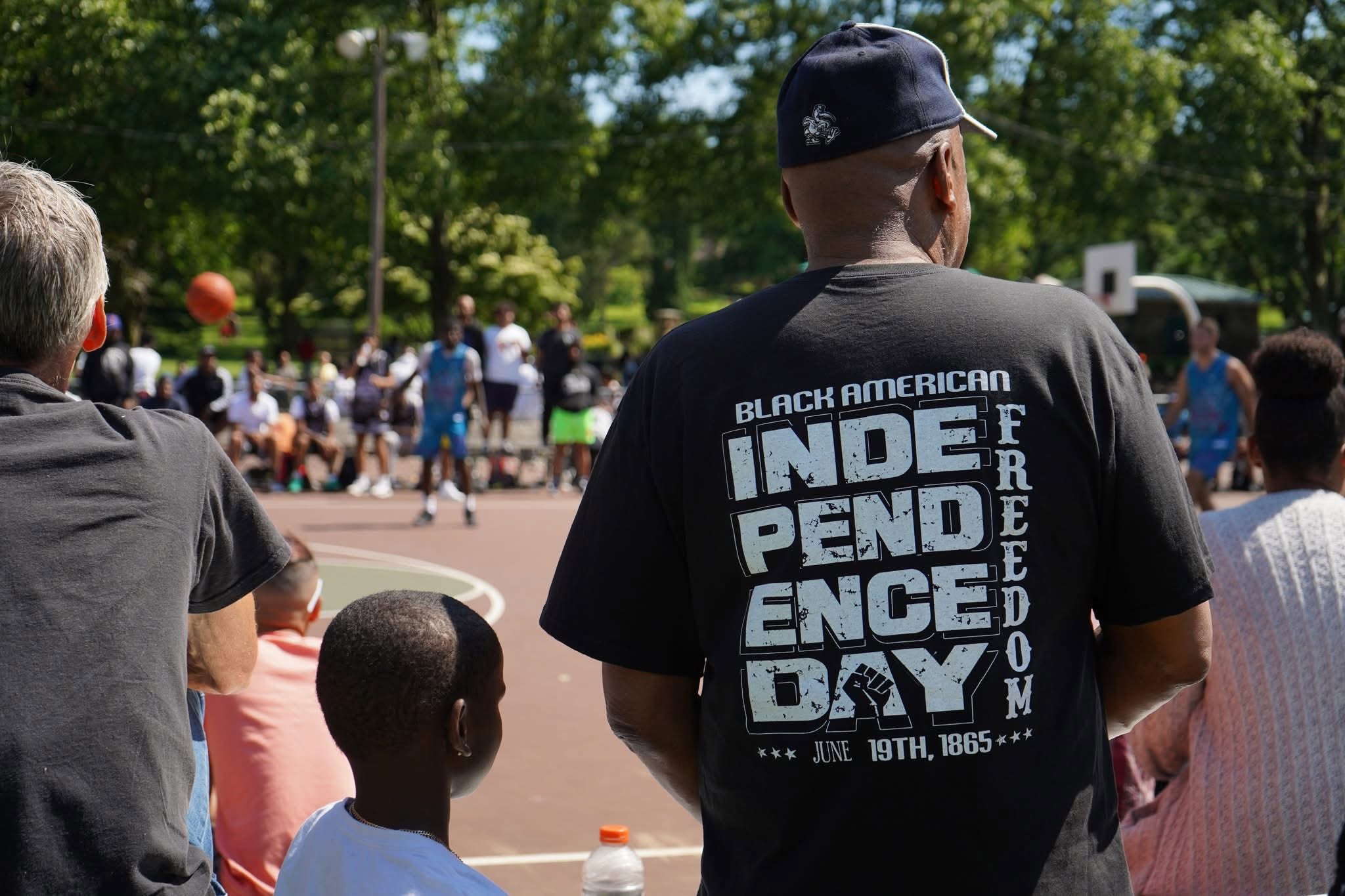 People watch an outdoor basketball game; one person wears a shirt saying 'Black American Independence Day'.