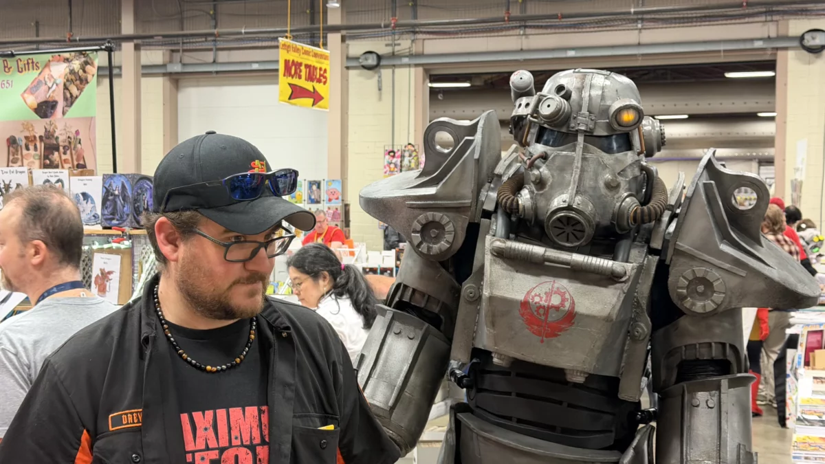 A man in a black shirt stands next to a person in a grey Fallout Power Armor costume.