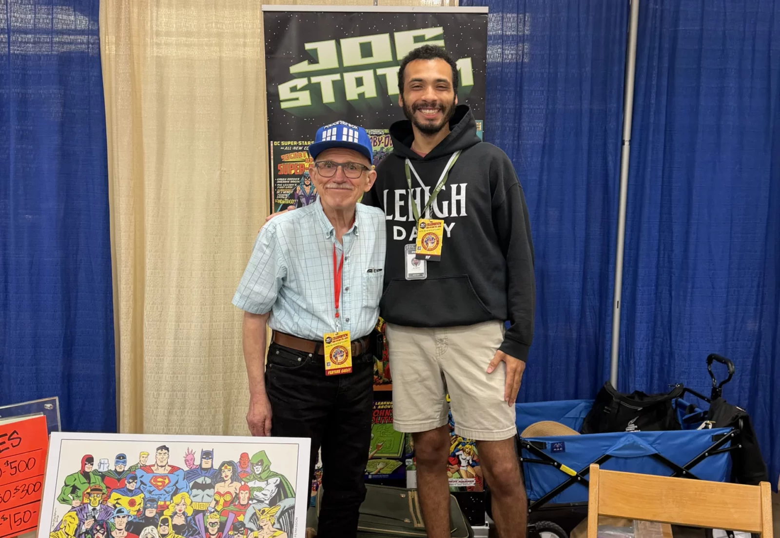 Reporter Jai Smith and comic artist Joe Staton stand smiling in front of a booth banner.