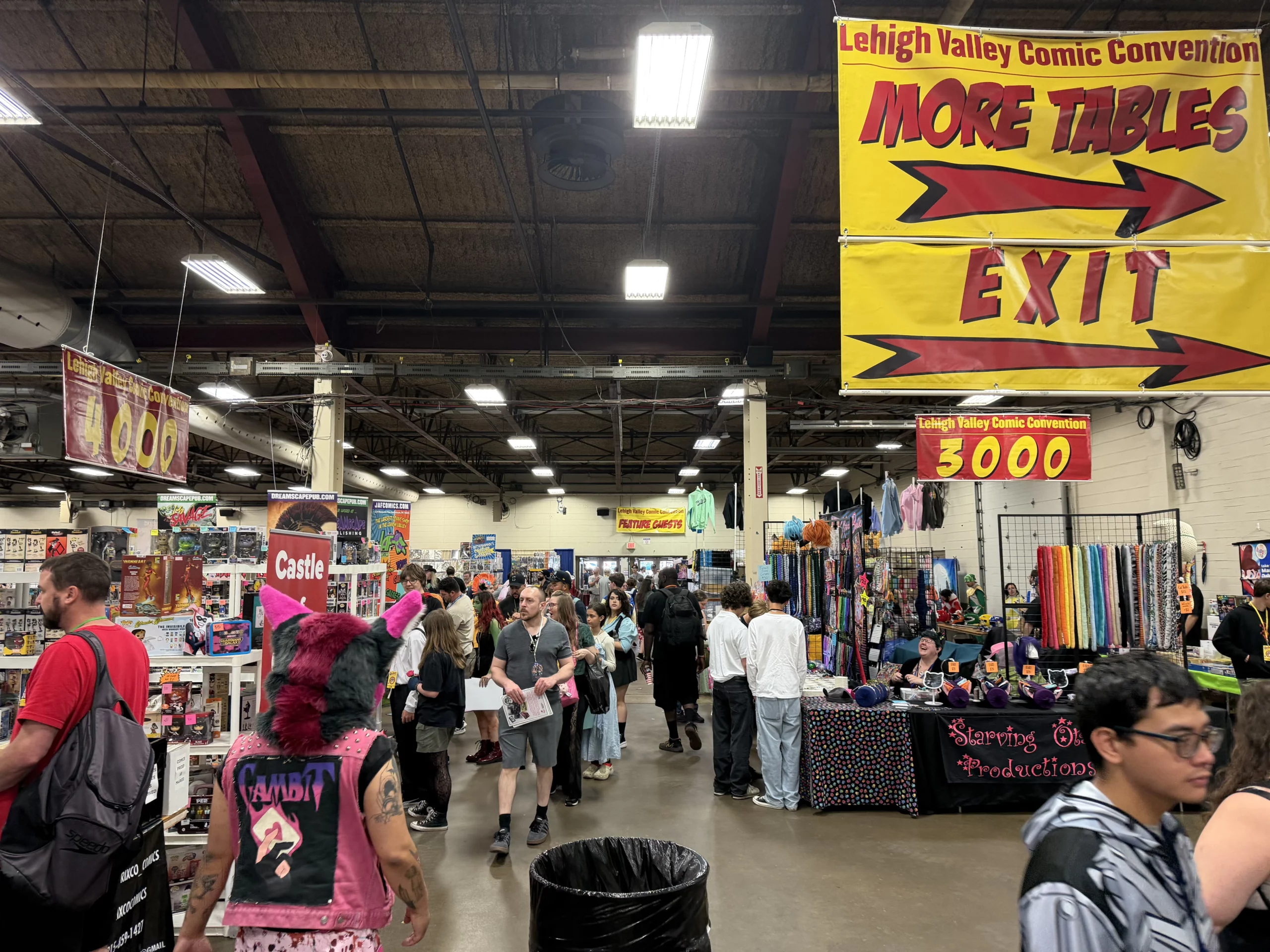 Crowded convention hall with attendees browsing vendor tables displaying comics, toys, and crafts.