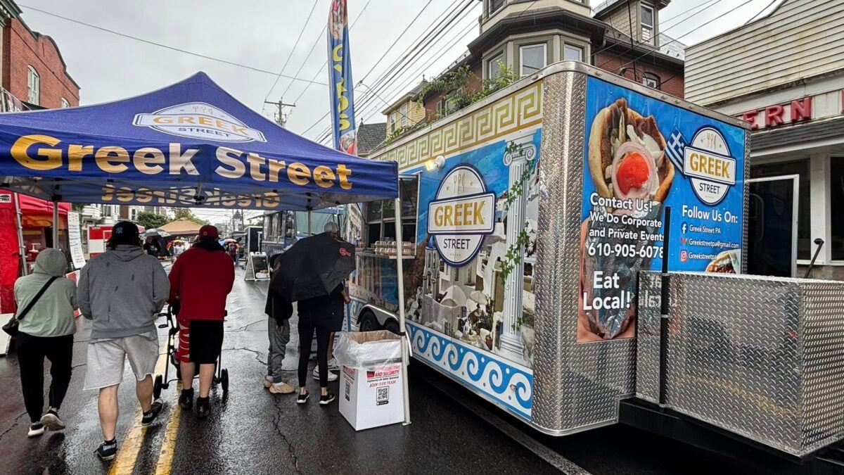 People walk on a wet street past a Greek Street food truck and blue vendor tent.
