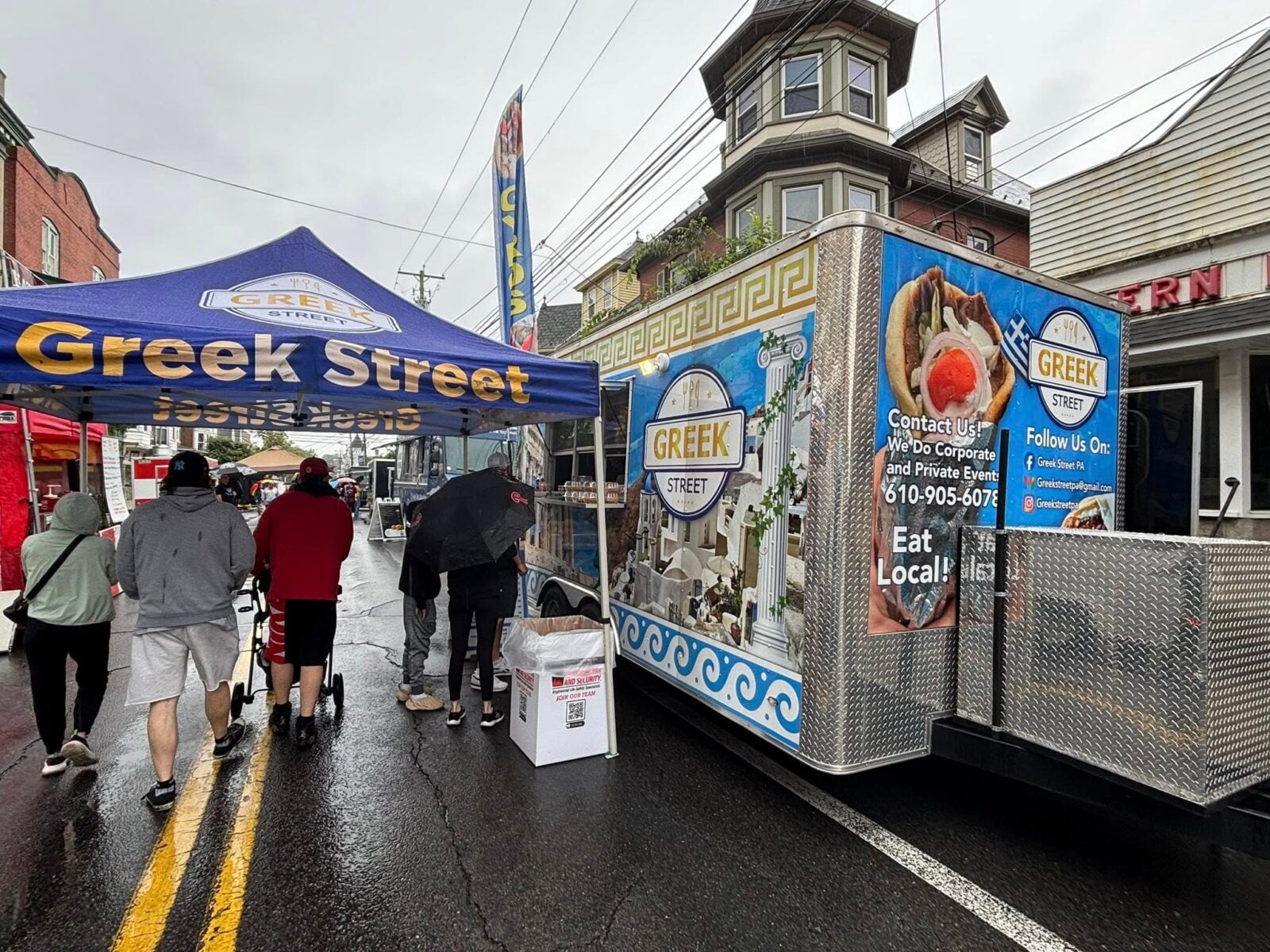 People walk on a wet street past a Greek Street food truck and blue vendor tent.