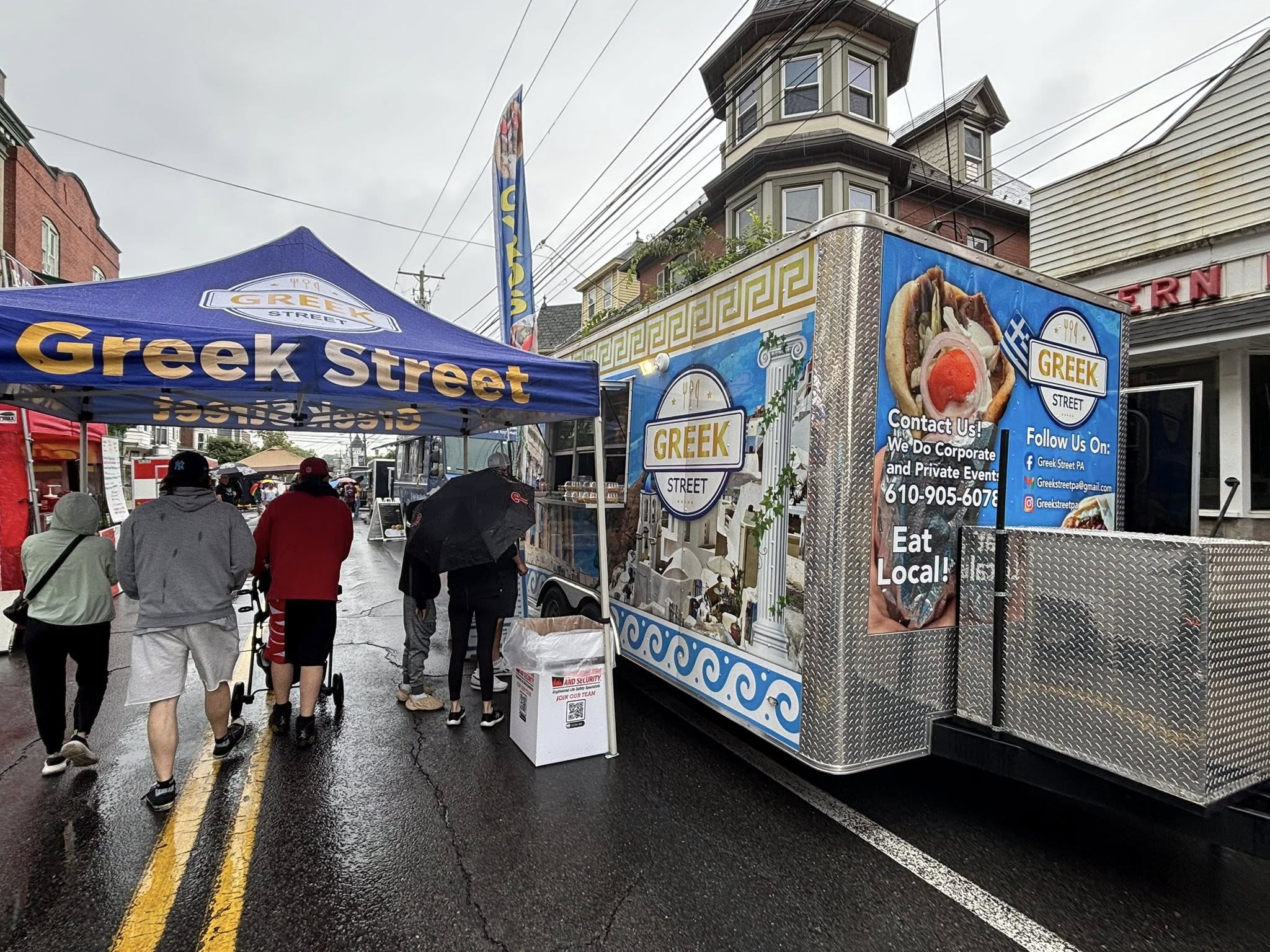 People walk on a wet street past a Greek Street food truck and blue vendor tent.