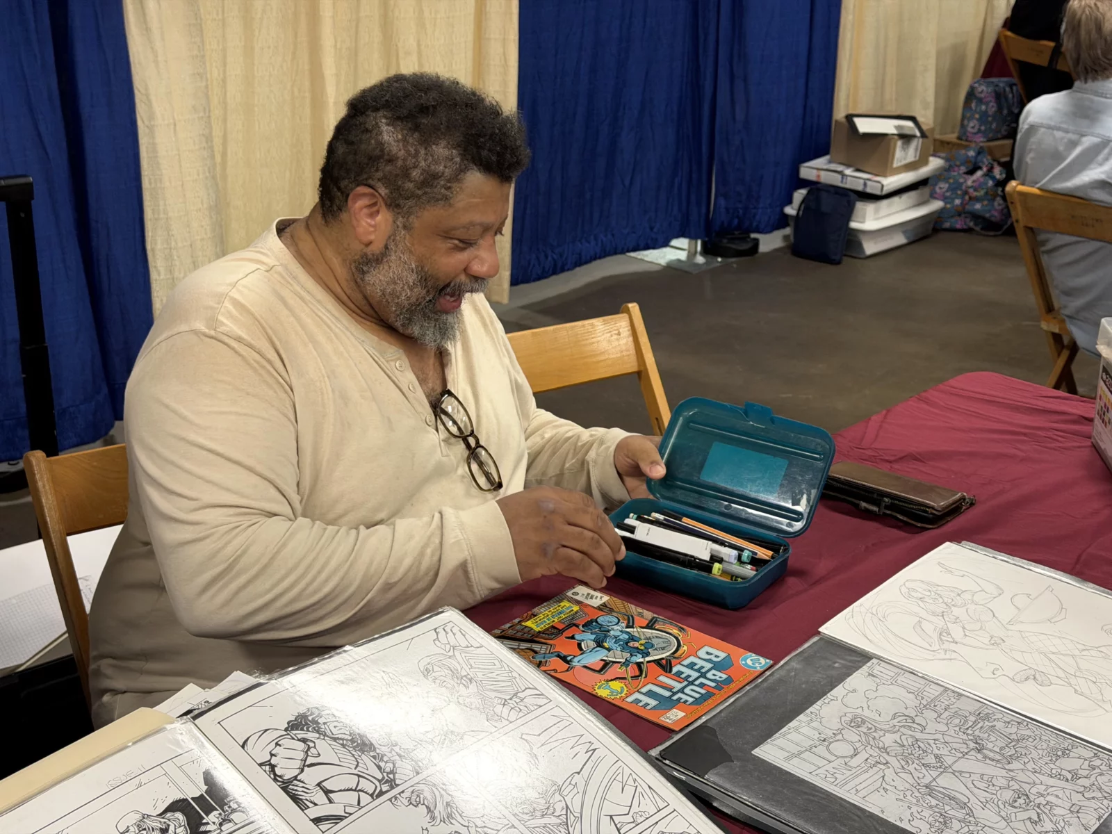 Bearded man in light shirt looks into teal pencil case with comic art on table.